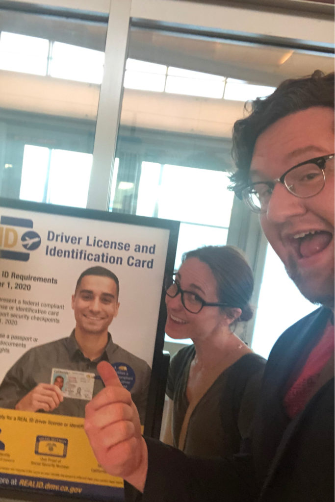 Casual photo of two people grinning at a California DMV poster about Real ID requirements